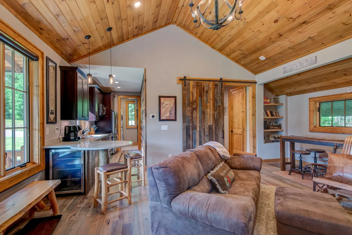 An open-plan space showing the seamless flow between the living room, kitchen, and dining area. The room features a wooden ceiling, a soft couch, and a dining table with an adjacent kitchen, all bathed in natural light from nearby windows.