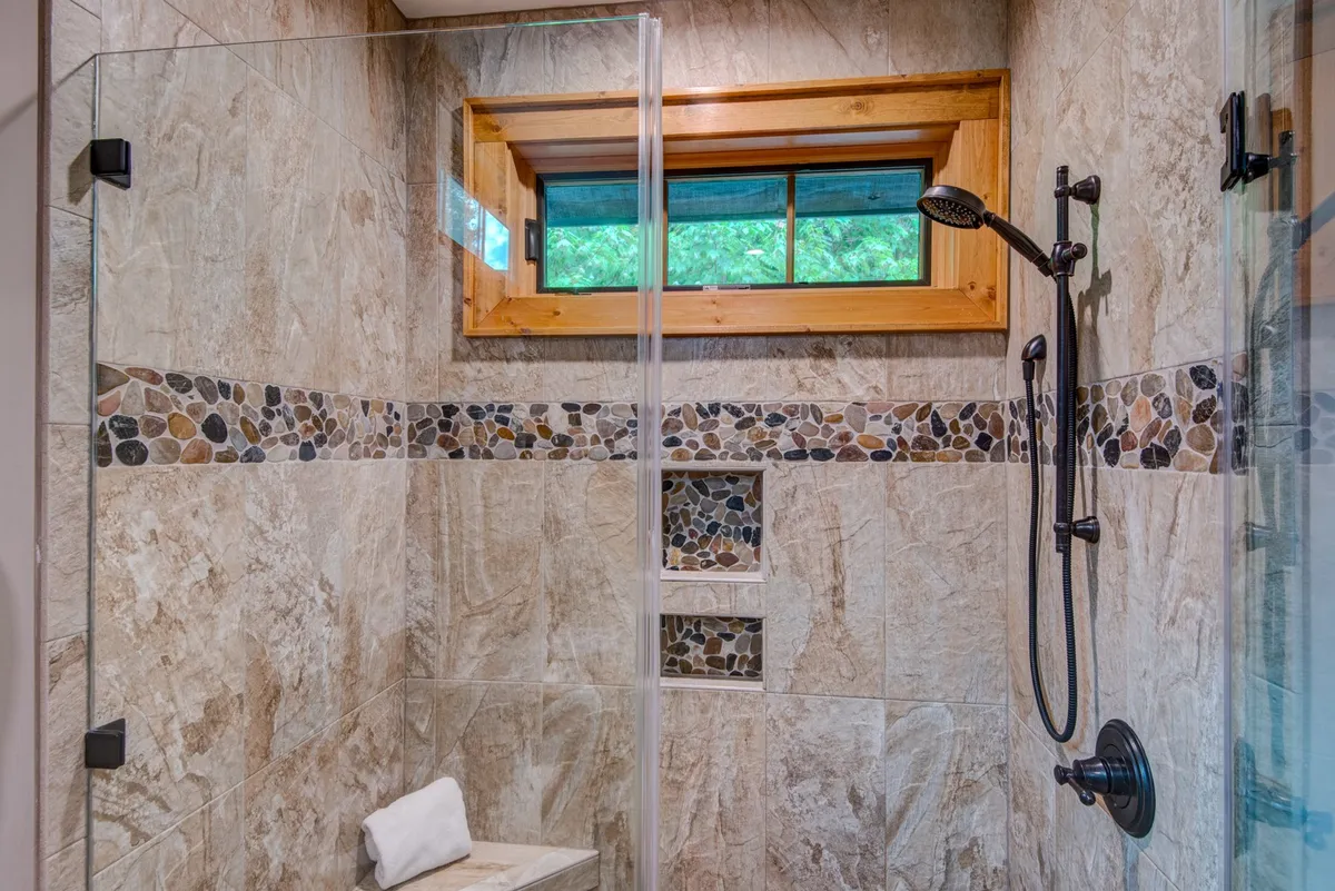 A stylish en-suite bathroom with a glass shower enclosure, natural stone tiles, and a wooden window frame. The scene radiates serenity, suggesting a refreshing shower experience after a day of adventure.