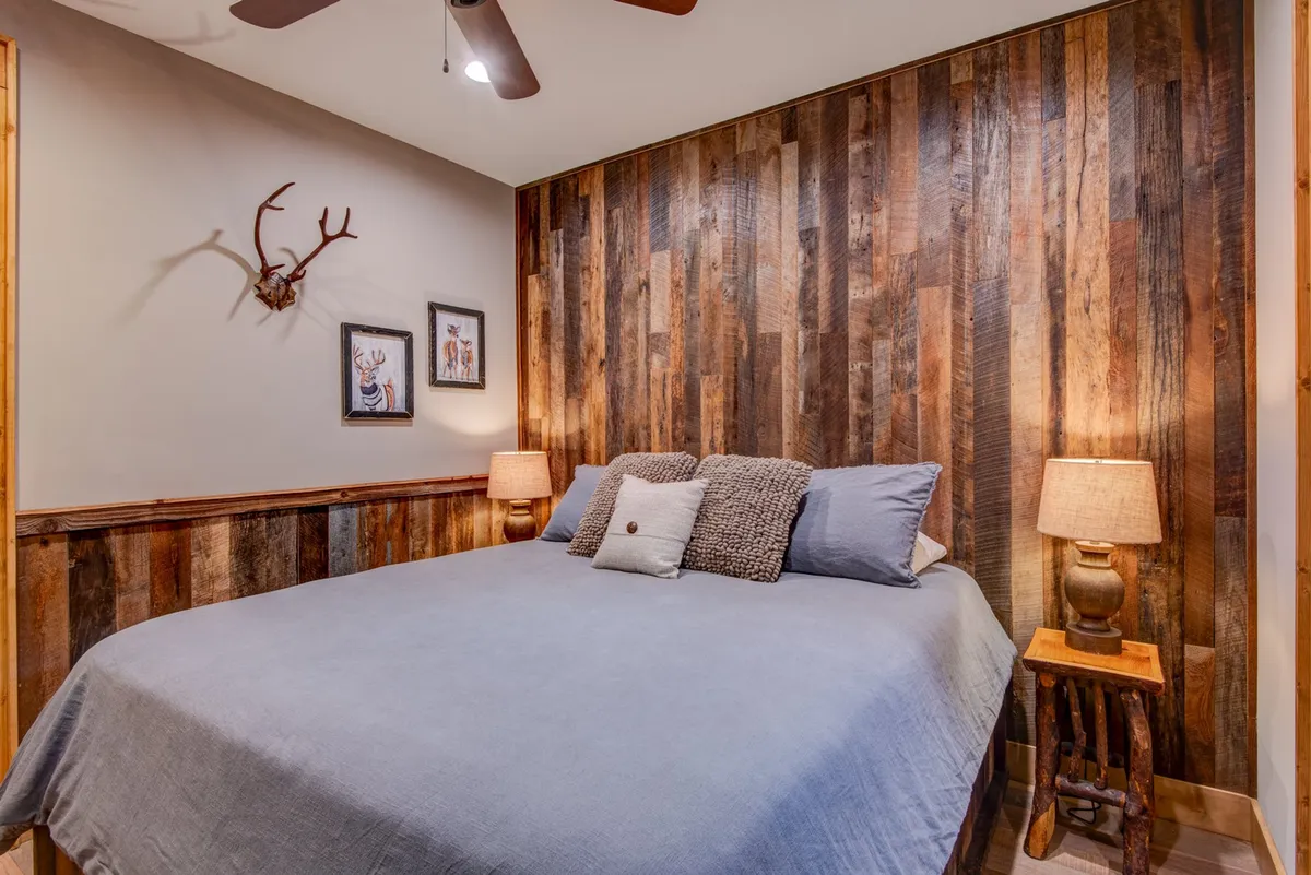 A warmly lit king bedroom featuring a wooden accent wall, a plush bed with crisp linens, nightstands with lamps, framed art, and decorative antlers. Soft natural light pours through a window, enhancing the inviting atmosphere.