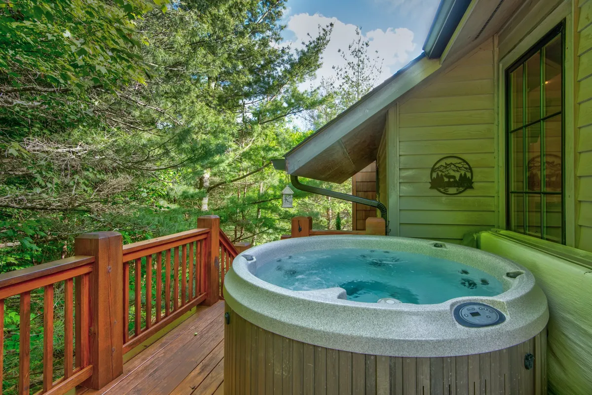 A serene scene of a bubbling hot tub set on a wooden deck surrounded by tall trees. Friends or family are relaxing in the hot tub, enjoying unobstructed views of the forested mountain setting. Soft evening light suggests an ideal moment for stargazing or unwinding after an active day outdoors.