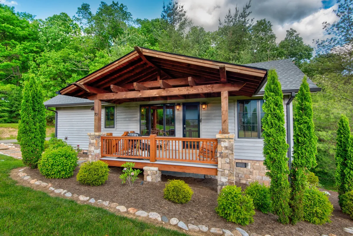 This exterior image features the front porch with sturdy stone columns, surrounded by lush greenery. The scene suggests a welcoming, communal space ideal for relaxing or socializing, with a backdrop of natural beauty. The sunset light adds an inviting warmth to the environment.