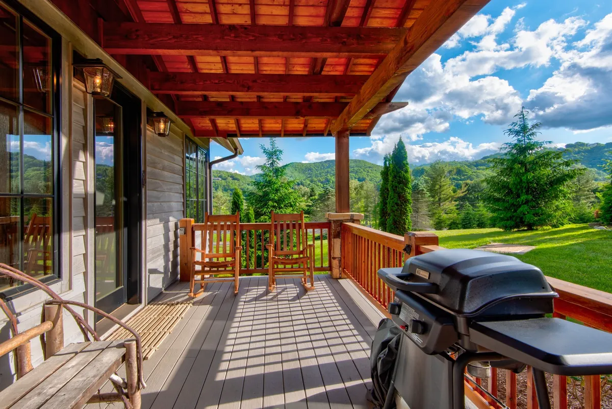 A view of a wooden deck featuring rustic rocking chairs, a grill for outdoor meals, and sweeping mountain vistas. The scene is set for either a quiet morning or a lively outdoor gathering, with ample space for pets and people alike.