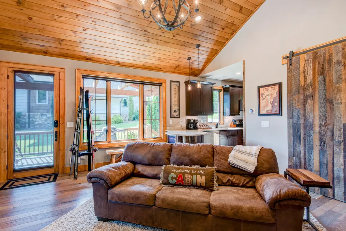 A welcoming, open living space with a leather sofa, exposed wooden beams, and a kitchen area, all illuminated by sunlight streaming through large windows. The design hints at easy access to outdoor spaces and social, family-friendly gatherings.