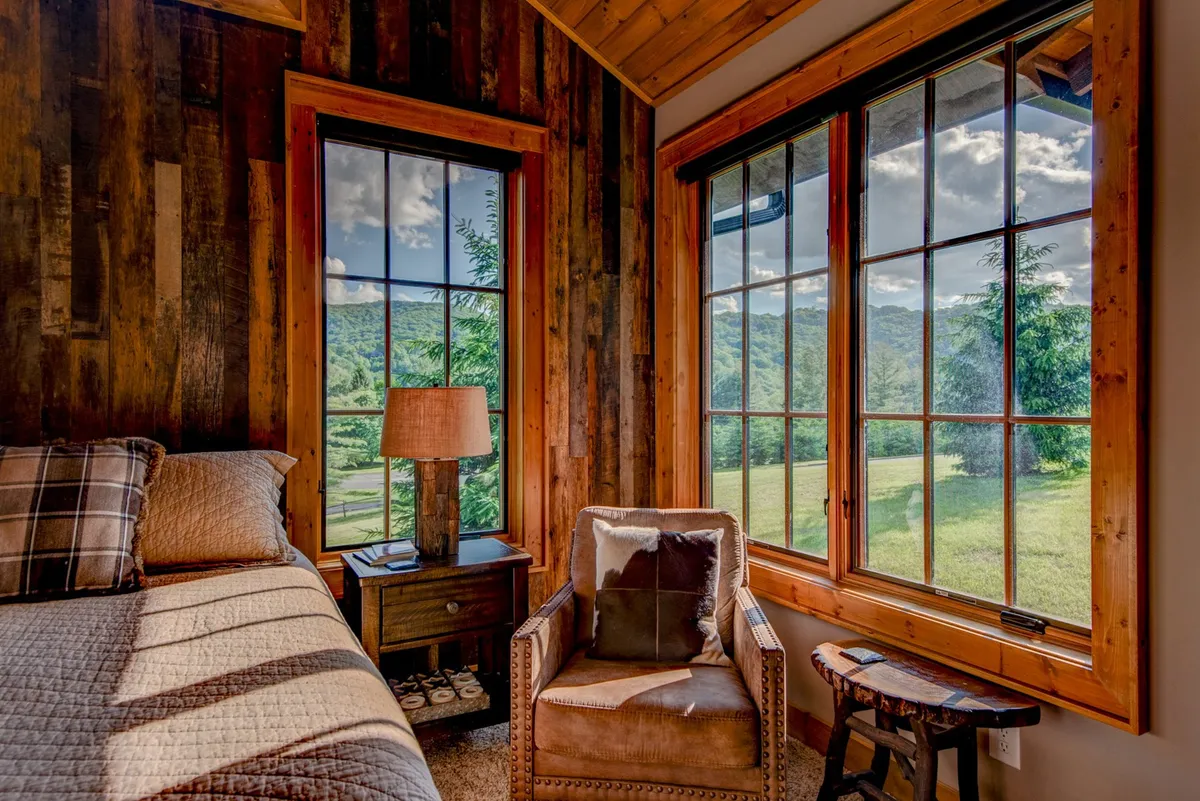 A sunlit bedroom with a plush bed adorned in textured bedding, flanked by two large windows. Natural light streams in, inviting guests to relax with a morning coffee or unwind in the comfortable chair beside the bed. Warm wood tones and thoughtful details create an intimate, restful atmosphere.