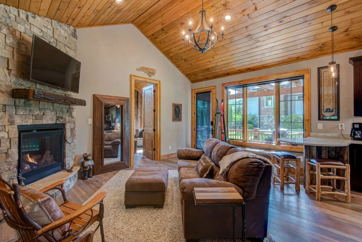An inviting living area with a stone fireplace, comfortable seating, and prominent, wide windows that frame mountain views. The wood-beamed ceiling and natural materials reinforce the cabin’s location within a mountain setting.